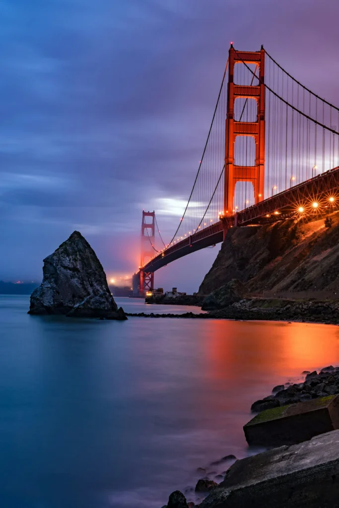 Illuminated Golden Gate Bridge tower next to a large offshore rock formation at twilight