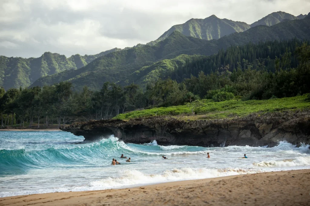 People swimming in rolling ocean waves next to a rocky cliff and lush green mountains