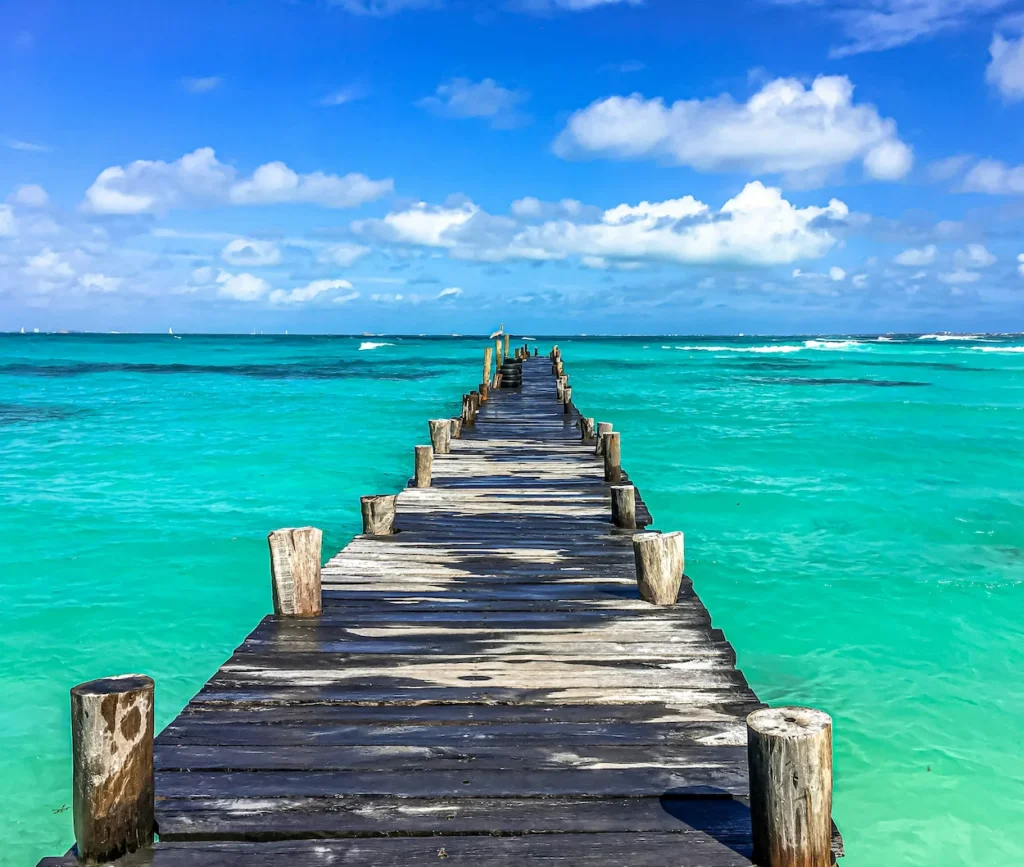 A weathered wooden pier stretching directly into bright turquoise ocean water under a cloudy blue sky