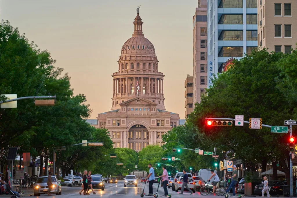 Texas State Capitol dome centered at the end of a tree-lined city street with pedestrians and traffic