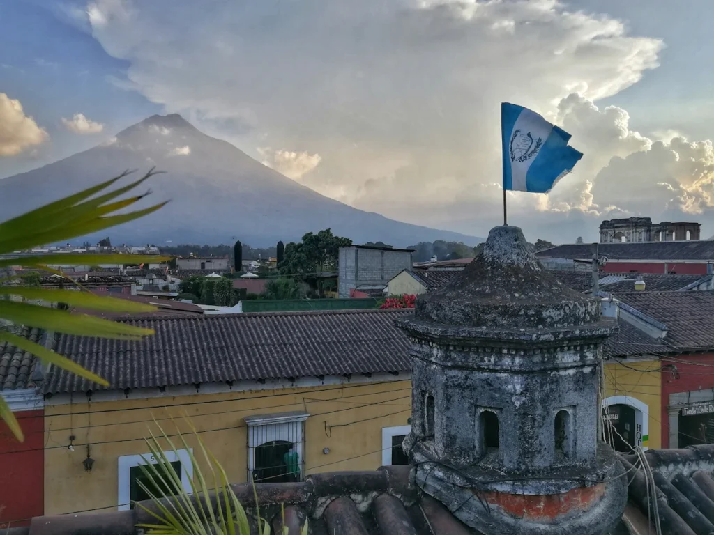 The flag of Guatemala flying on a stone cupola overlooking rooftops and a large volcano