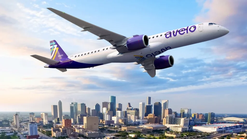 Avelo Airlines airplane flying low at dusk with an illuminated city skyline visible below