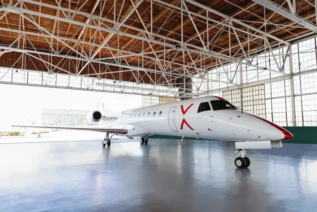 White JSX Embraer 145 passenger jet parked inside a spacious airplane hangar with a wooden truss ceiling