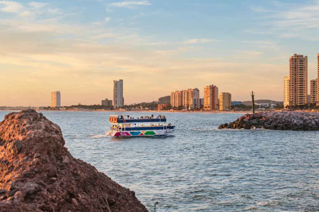 A colorful double-decker tour boat cruising on the ocean with the Mazatlán skyline in the background
