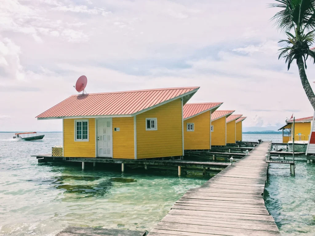 A row of bright yellow overwater bungalows with orange roofs lined up along a wooden boardwalk over clear water