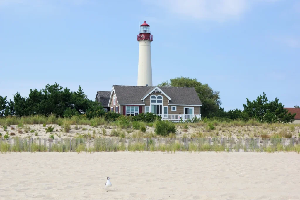 The Cape May Lighthouse rising behind a coastal house and a sandy beach under a blue sky