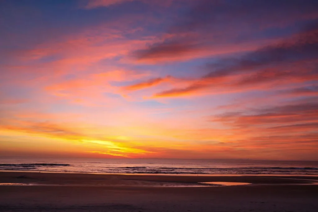A dramatic sunrise casting bright pink, orange, and purple hues across clouds over a quiet beach