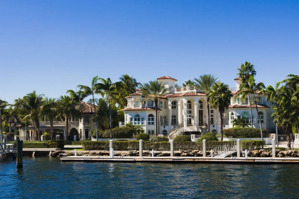 Large multi-story white luxury mansion with palm trees and a private boat dock on the waterfront
