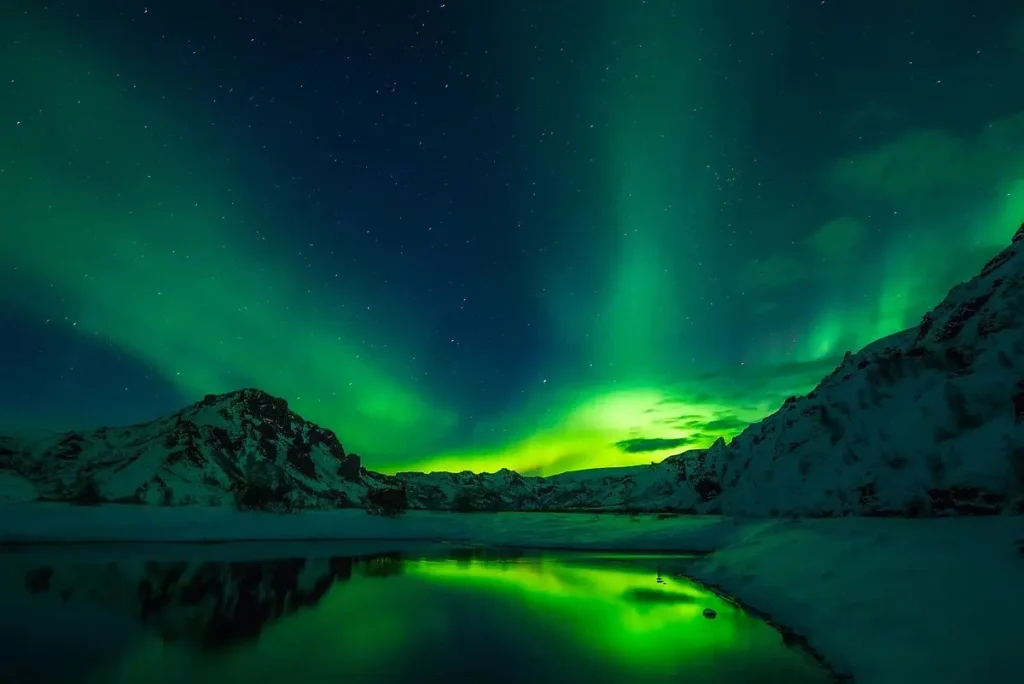 The Northern Lights glow green over snowy mountains and a reflective lake in Iceland.