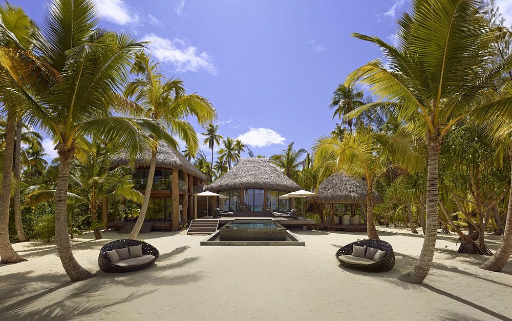The Brando resort villa with thatched roofs, pool, and palm trees on a sandy beach.
