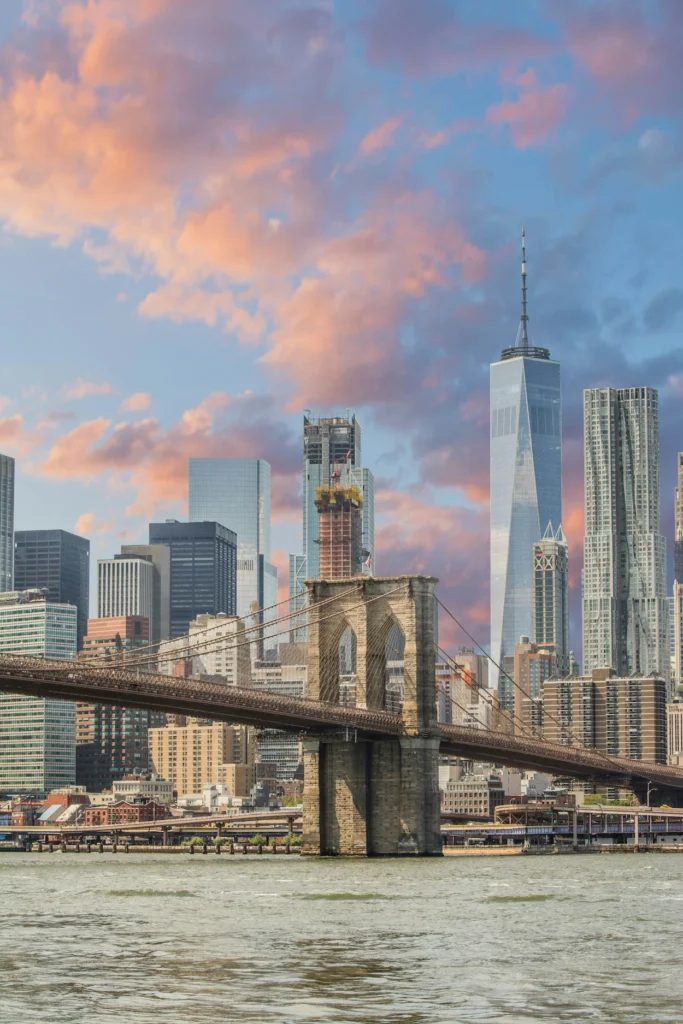 Brooklyn Bridge spanning the East River with One World Trade Center and Manhattan skyscrapers under a sunset sky