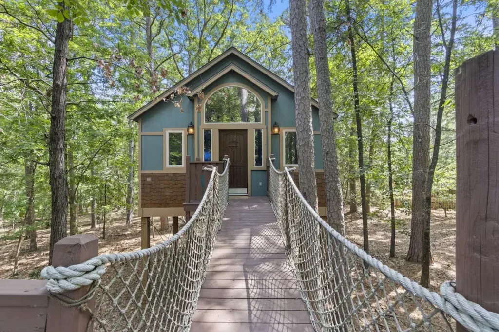 A wooden suspension bridge leading to a green and teal cabin surrounded by trees