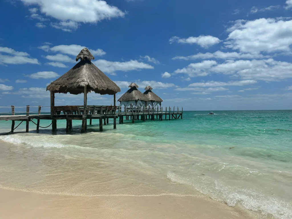 Wooden pier with thatched-roof palapas extending into turquoise ocean water from a sandy beach