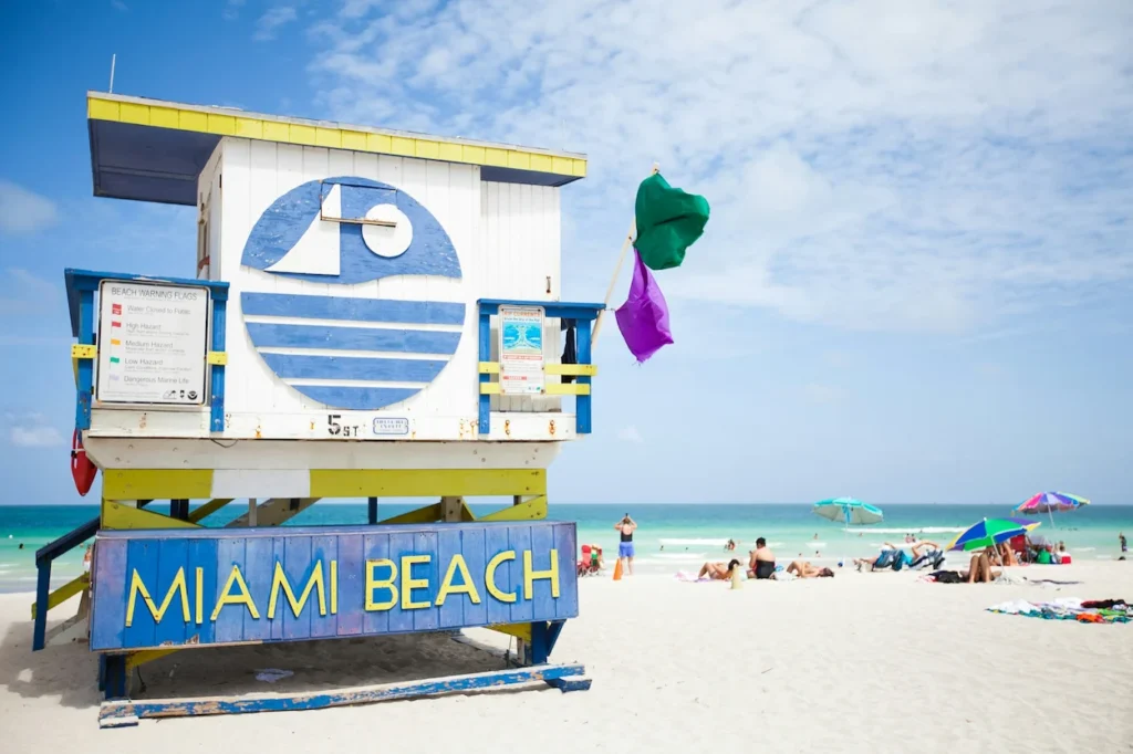 White, blue, and yellow 5th Street lifeguard tower on sandy beach with sunbathers and turquoise ocean water