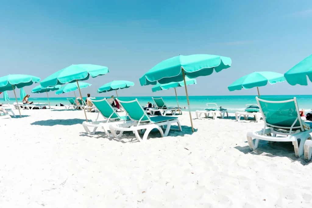 Turquoise umbrellas and matching lounge chairs arranged on bright white sand facing a calm teal ocean