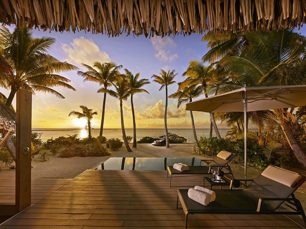 The Brando resort deck with lounge chairs, pool, and palm trees overlooking a sunset beach.