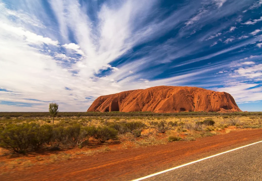Uluru monolith in the Australian outback desert seen from a road under a blue sky with wispy clouds
