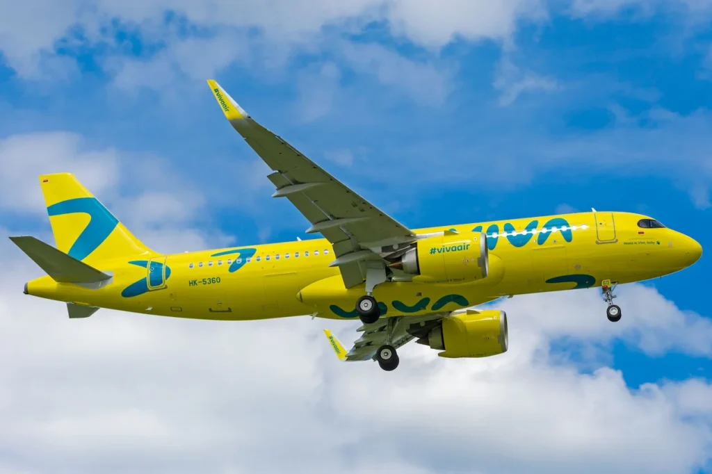 A bright yellow and blue Viva Air Airbus A320 passenger airplane flying through a cloudy blue sky