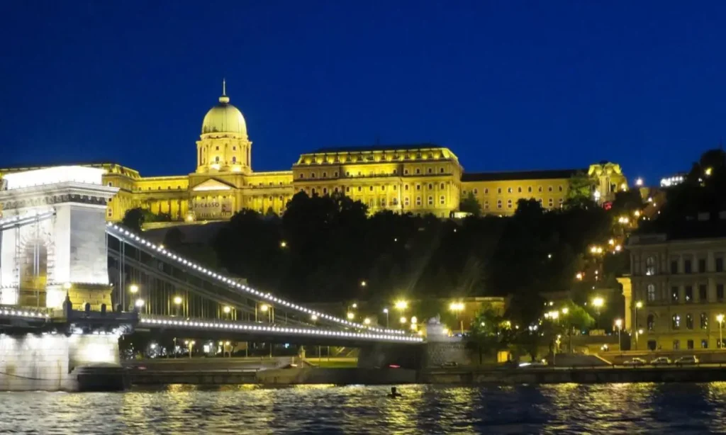 The Danube river flows beside a lit-up bridge and grand building at night.