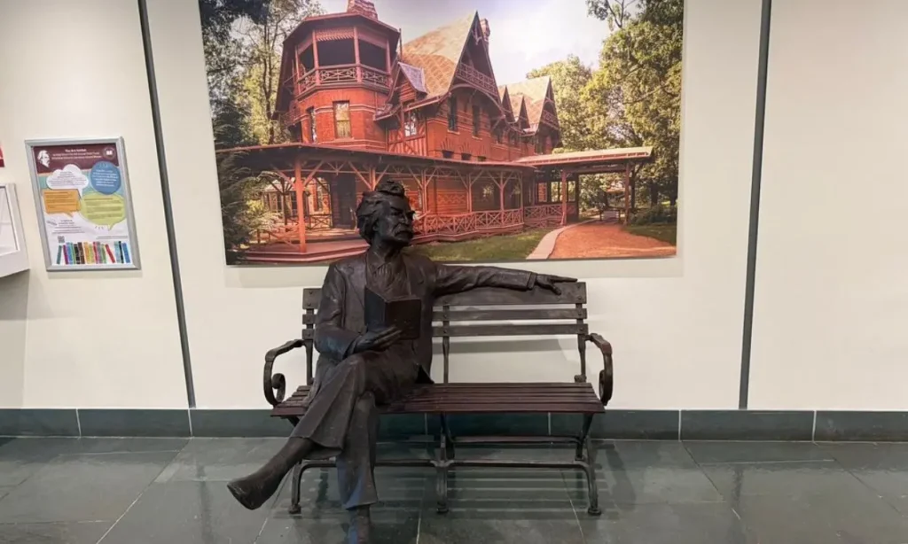 Bronze statue on a bench in front of a large photo of The Mark Twain House & Museum.