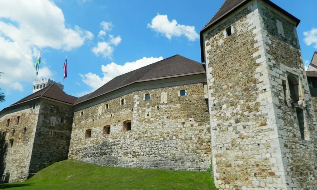 Ljubljana Castle has stone walls and towers sitting on a grassy hill under a bright sky.
