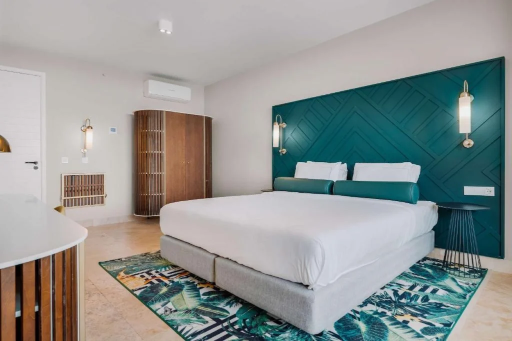 Modern hotel bedroom with a large bed, teal geometric accent wall, and tropical leaf area rug
