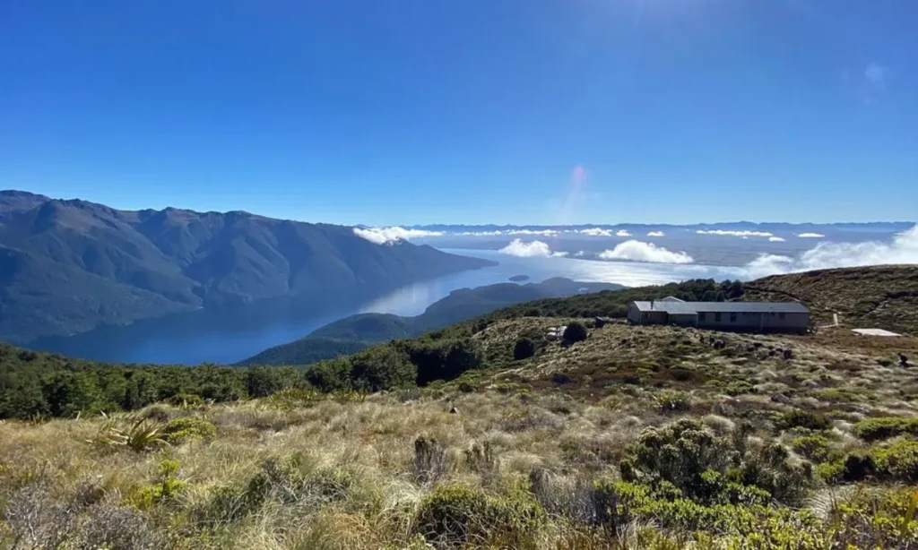 Scenic view of Fiordland National Park with mountains, a lake, and a hut under a clear blue sky.