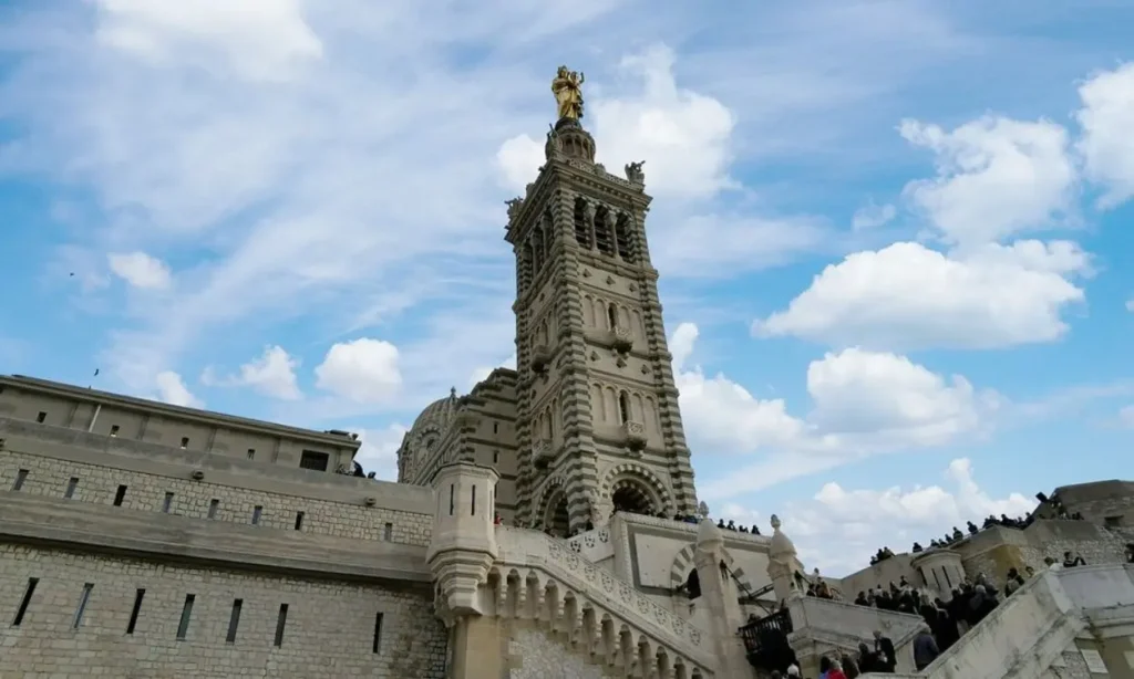 The tower of Basilique Notre-Dame de la Garde rises above its stone steps against a cloudy sky.