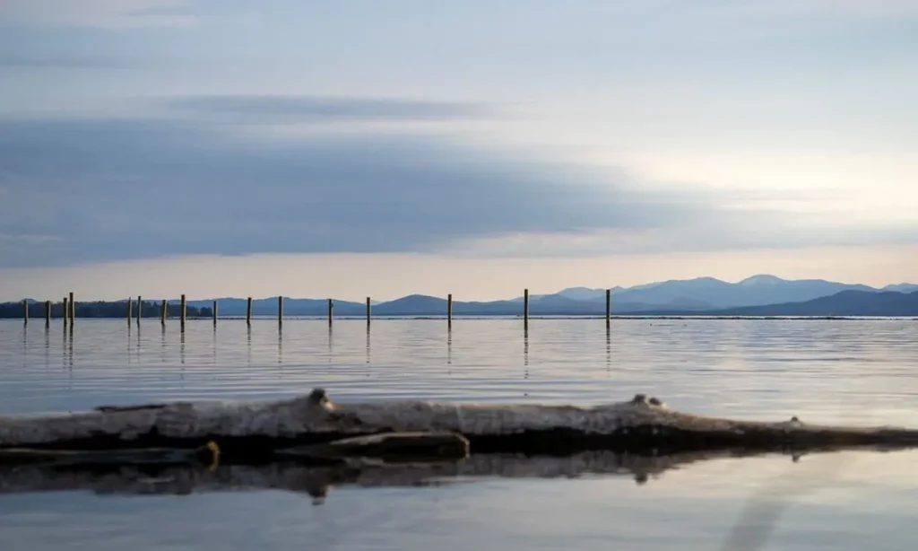 Calm water reflects the sky and mountains at Lake Champlain with wooden posts in the distance.