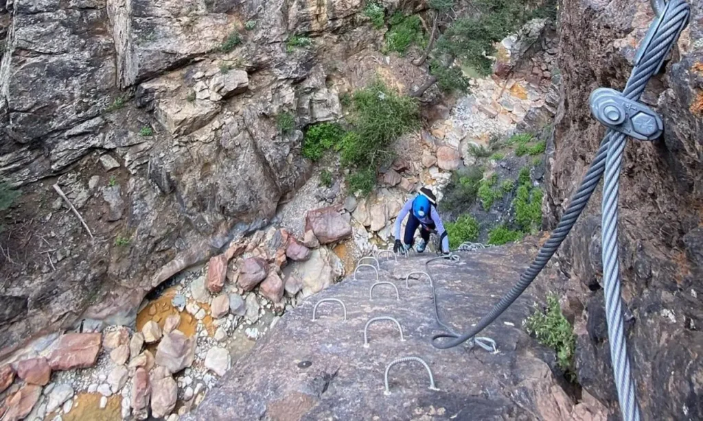 A climber ascends the Ouray Via Ferrata using metal rungs and cables on a rock wall.