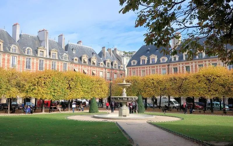 Park at Place des Vosges with symmetrical trees, fountain, and historic buildings in the background.