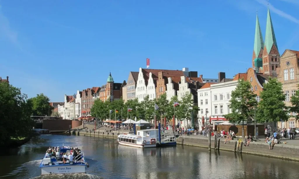 A boat cruises down the river with Lübeck’s historic buildings and church towers in the background.