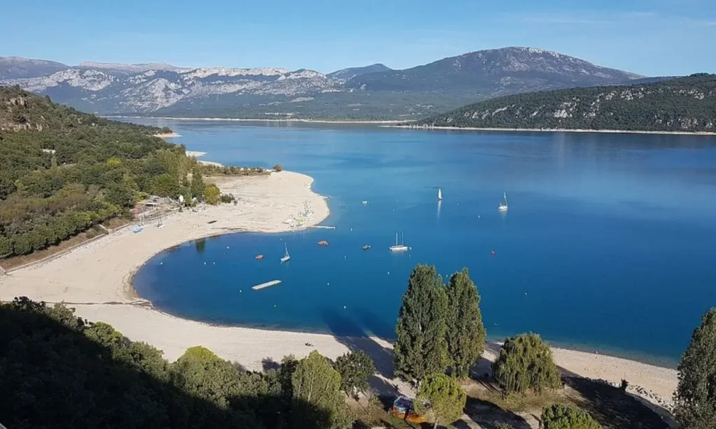 A sandy beach curves around the blue waters of Gorges du Verdon with mountains in the distance.