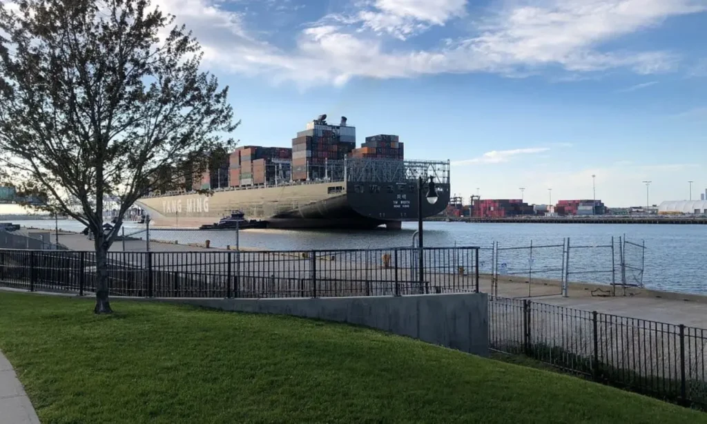 A large cargo ship sailing past The Hudson River Waterfront Walkway on a clear day.