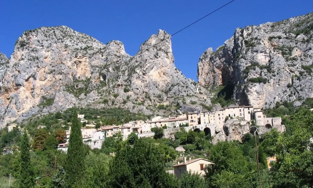 A row of houses in Luberon sits at the base of tall rocky mountains under a clear blue sky.
