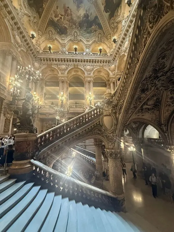 Grand staircase inside the Opéra Garnier with ornate chandeliers, sculptures, and a painted ceiling.