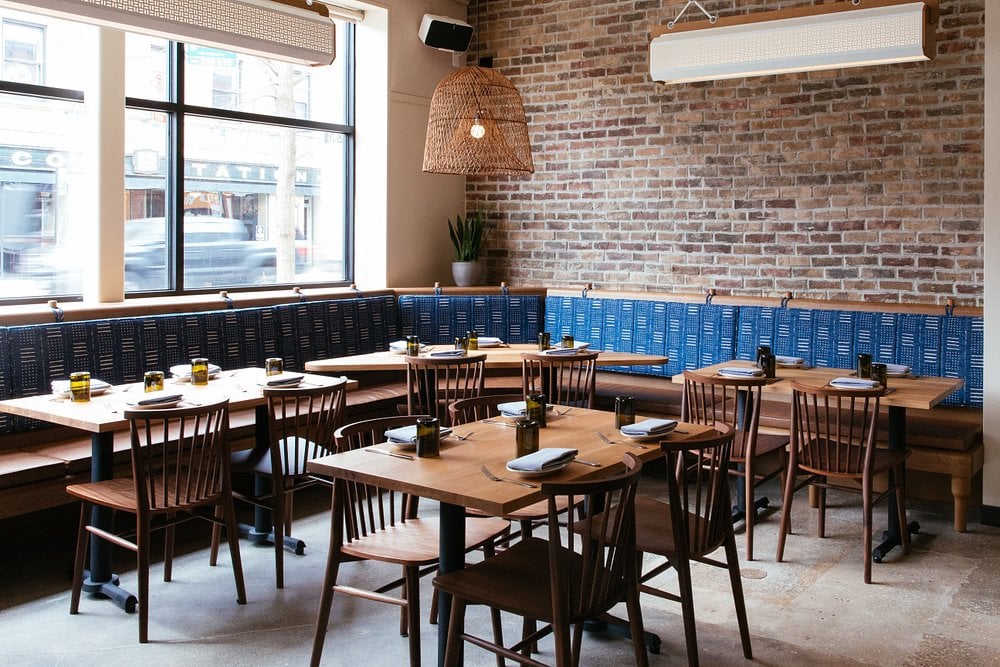 Cozy and modern dining area at Galit with wooden tables, blue bench seating, and brick walls.