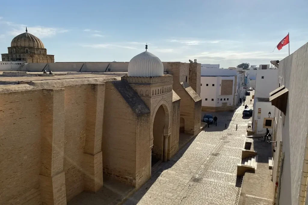 A street view in Kairouan with the Great Mosque’s domes and a Tunisian flag in the distance.