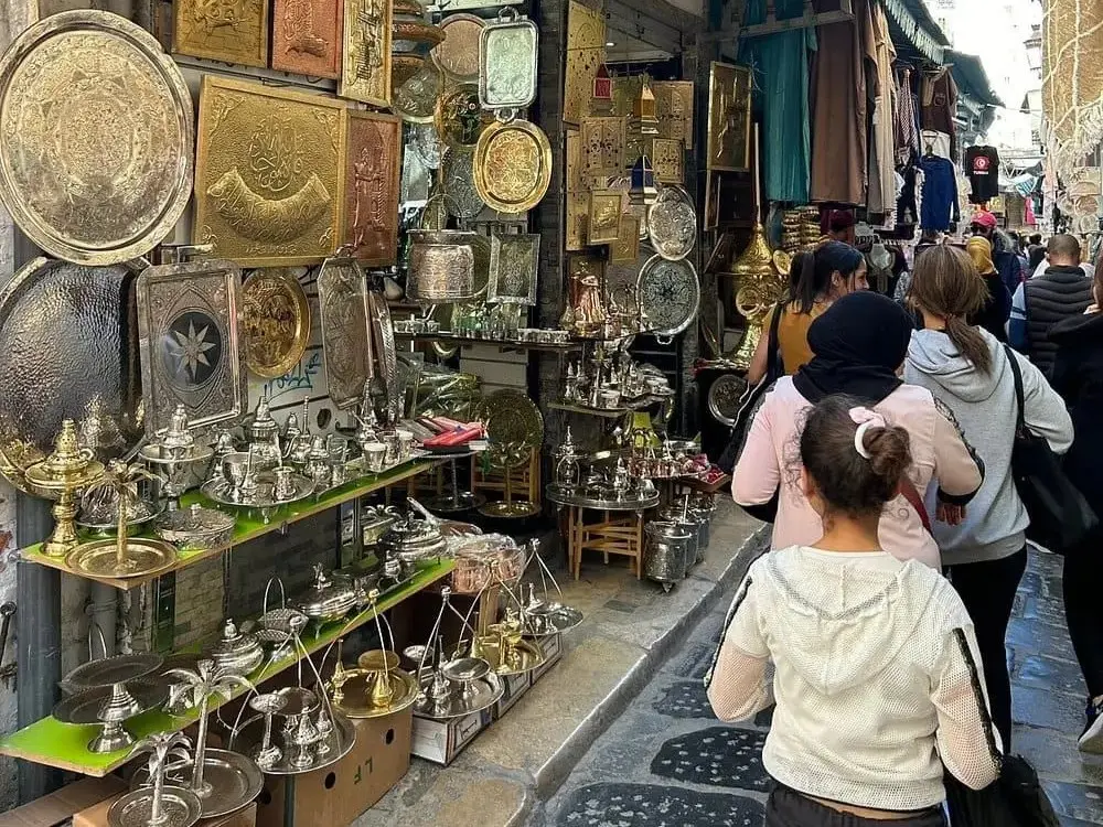 People walking through the bustling Medina of Tunis with shops selling brassware and traditional crafts.