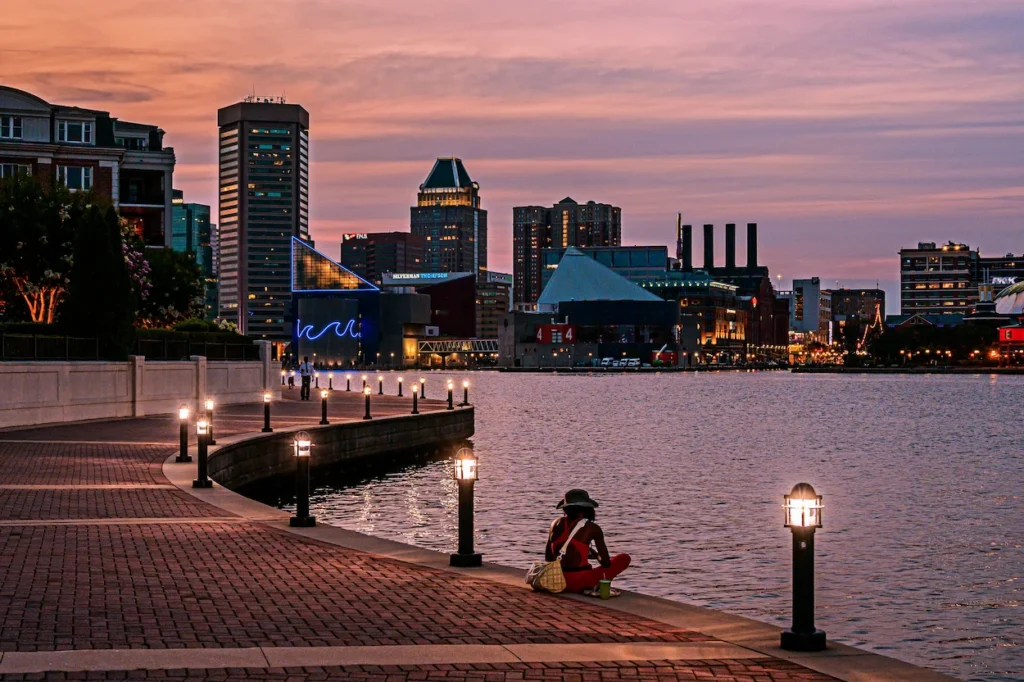 Person sitting on a brick promenade pathway by the water with the Baltimore Inner Harbor skyline at sunset in Maryland