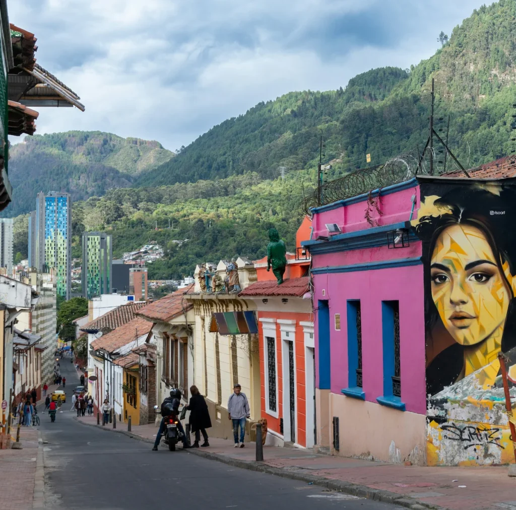 A steep street in Bogota lined with colorful houses and a large face mural against green mountains