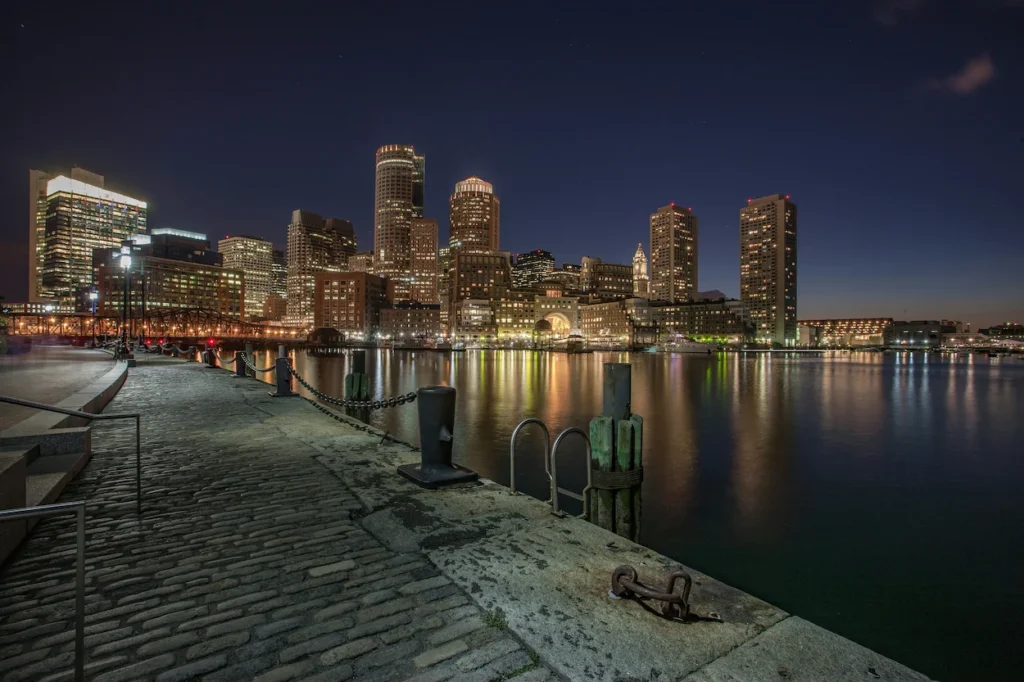 Boston skyline lit up at night across the water from a stone harborfront walkway