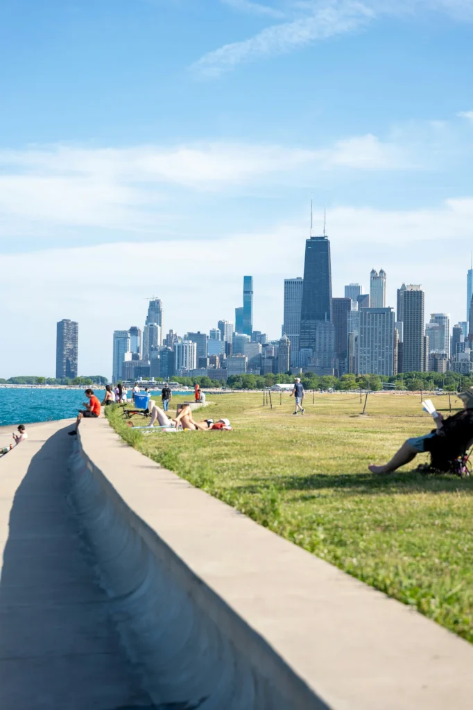 People relaxing on a grassy shore by a concrete seawall with the downtown Chicago skyline across Lake Michigan