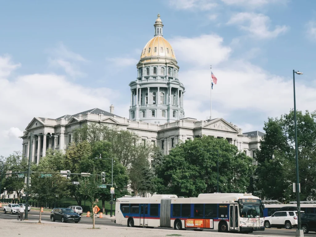 Colorado State Capitol building with its golden dome behind green trees and a street in Denver