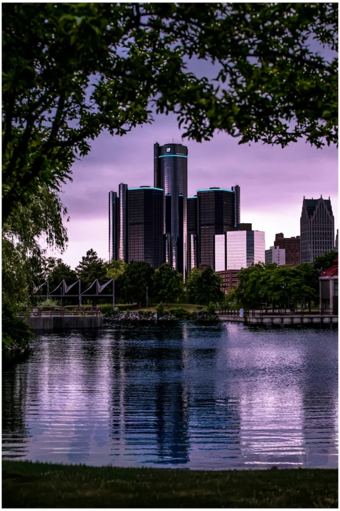 Detroit Renaissance Center towers reflecting on a river at twilight, framed by dark green tree branches