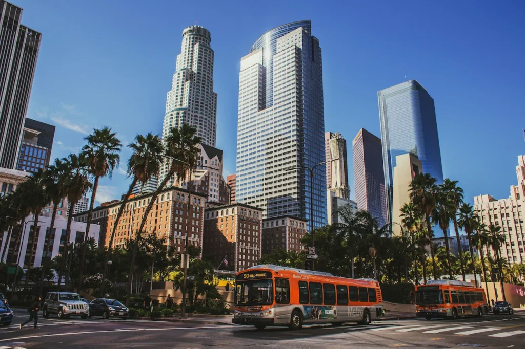 An orange LA Metro bus driving past palm trees and skyscrapers near Pershing Square