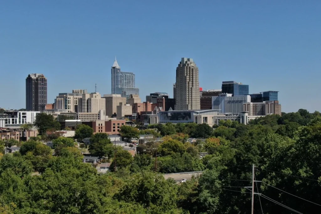 Downtown skyline of Raleigh rising above a dense forest of green trees under a clear blue sky