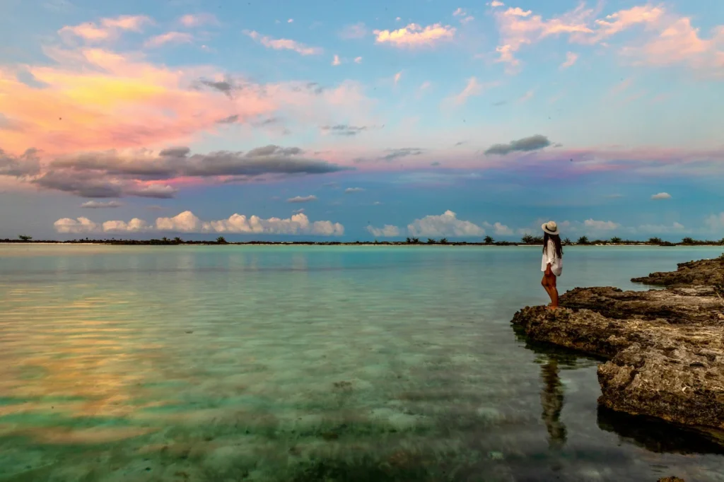 Woman in a sun hat on rocky shore overlooking a calm pink sunset lagoon in Eleuthera, Bahamas