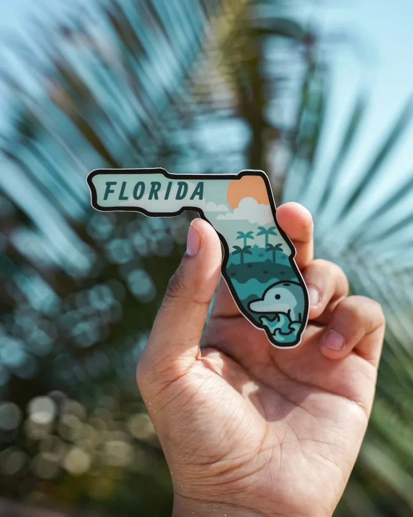 Hand holding a Florida map sticker featuring a manatee and palm trees against blurry palm leaves