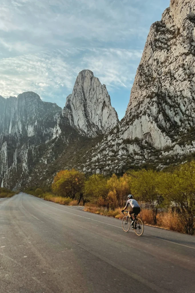 Cyclist riding down a paved mountain road flanked by towering limestone peaks under a pale blue sky
